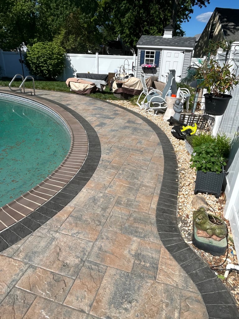 Backyard pool area with curved patio deck, lounge chairs, white storage shed, landscaping, and trees under blue sky