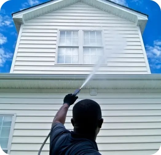 Person pressure washing white vinyl siding on a two-story house under a blue sky