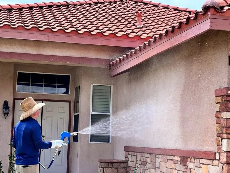Person in blue jacket and straw hat power washing a house exterior wall with spray gun