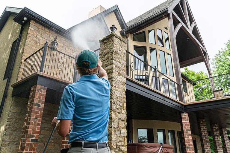 Worker in blue shirt and hard hat pressure washing the brick exterior of a large two-story house