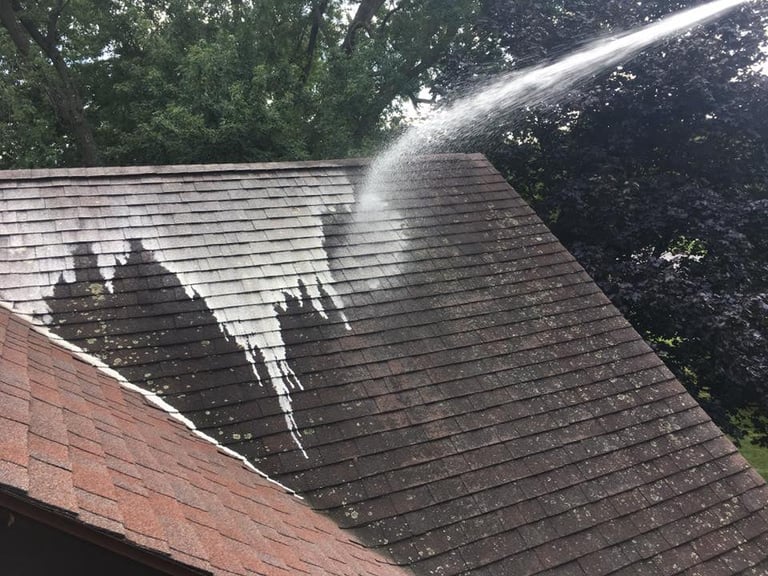 Roof being pressure washed with water spray, showing water stream cleaning dark shingled roof surface