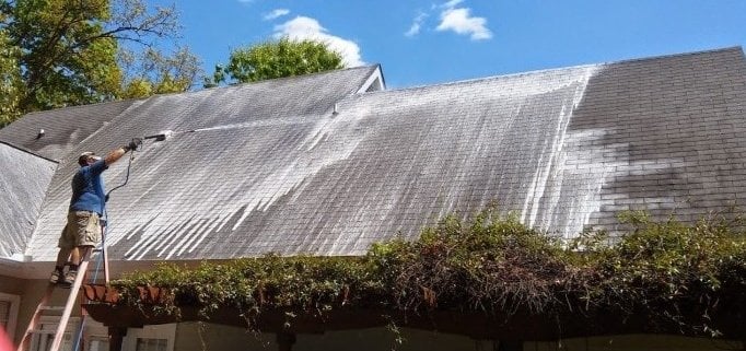 Worker using pressure washer to clean moss and algae off a steep residential roof with clear sky in background