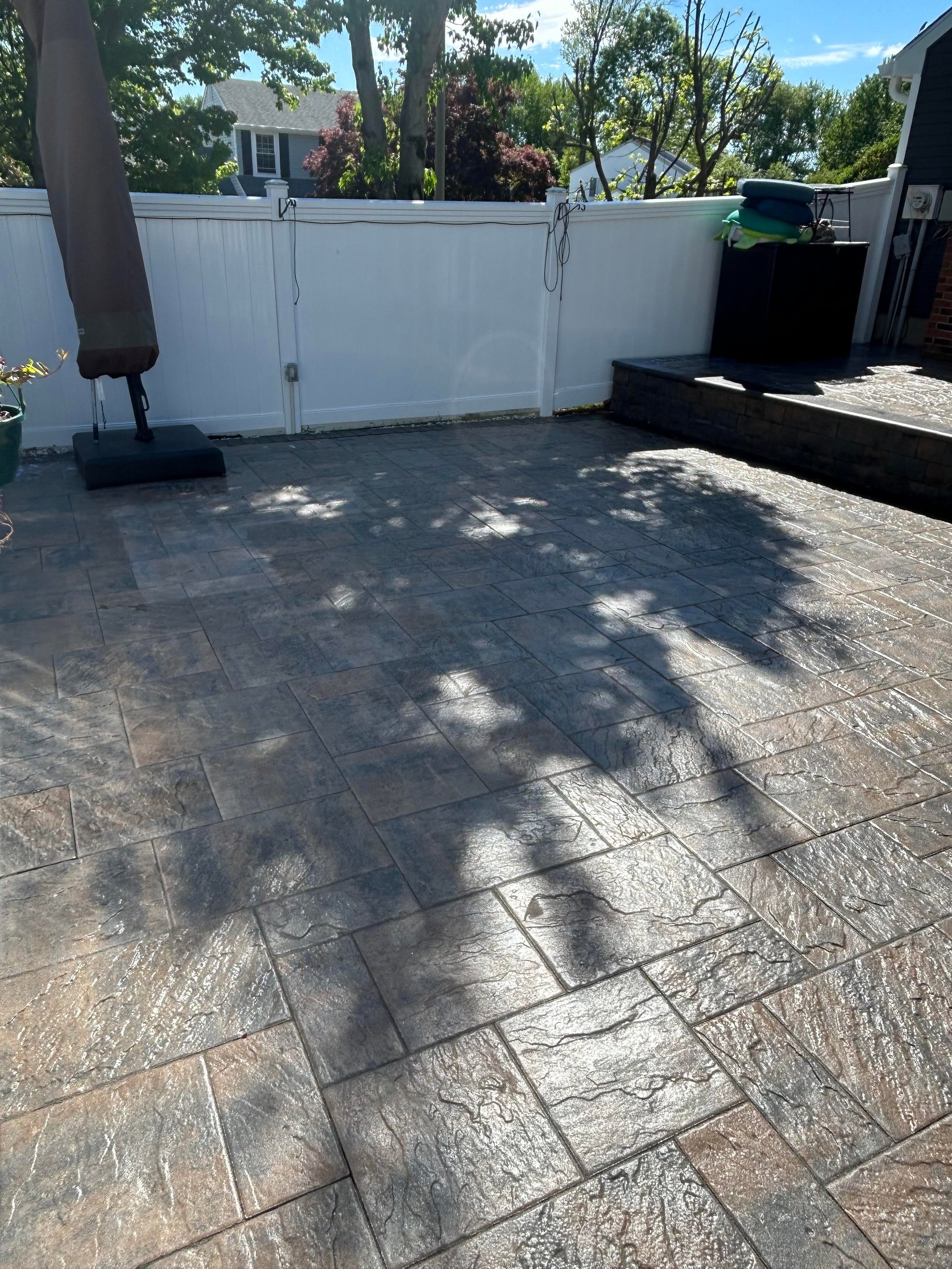 Stamped concrete patio with white fence, umbrella base, and trees in background on sunny day