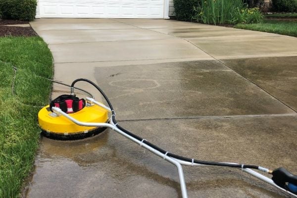 Yellow surface cleaner attached to hose on wet driveway with garage and lawn in background