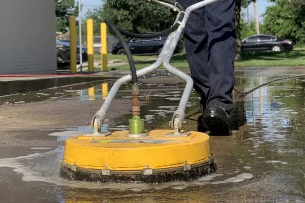 Worker operating yellow surface cleaning equipment on wet pavement during pressure washing or cleaning service