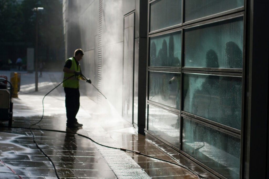 Worker in yellow safety vest pressure washing a storefront building facade with water spray